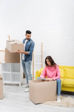Woman holding book near muslim boyfriend with carton box at home 