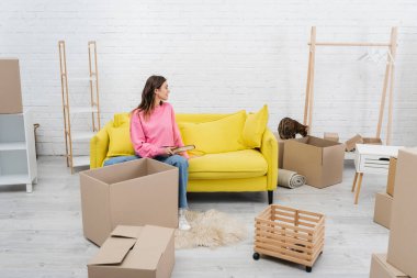 Side view of woman holding book near bengal cat and carton boxes at home 