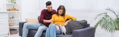 Positive multiethnic couple looing at sphynx cat on couch at home, banner  clipart