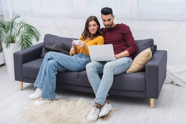 Smiling woman holding cup near muslim boyfriend with laptop on couch at home 