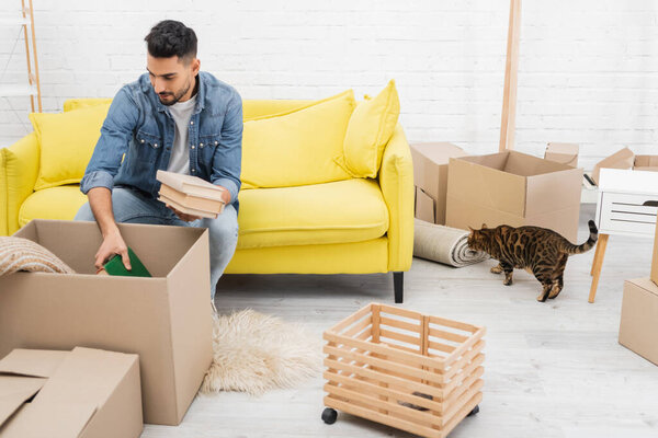 Arabian man holding book while unpacking boxes near bengal cat at home 