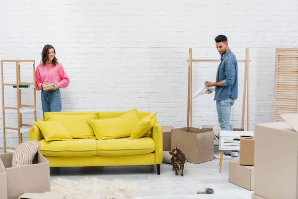 Smiling Multiethnic Couple Looking Bengal Cat While Unpacking Boxes Home — Stock Photo, Image