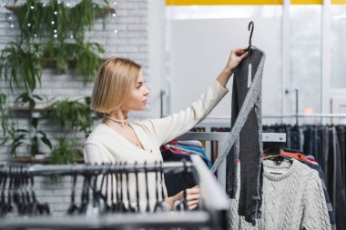 Side view of young woman holding clothes near hangers in vintage shop 