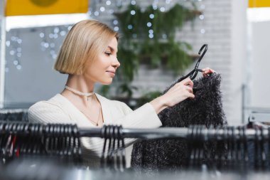 Side view of young blonde woman choosing clothes in second hand 