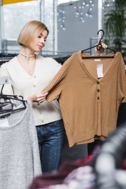 Young woman holding hanger with blouse in second hand 