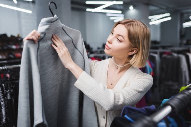 Young blonde woman touching sweatshirt on hanger in second hand 