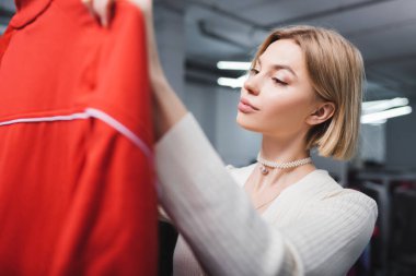 Young blonde woman holding blurred clothes in second hand 