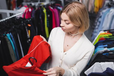 High angle view of young blonde woman looking at clothes near blurred hangers in vintage shop 