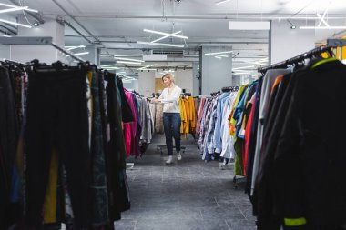 Side view of smiling woman walking near clothes in second hand 