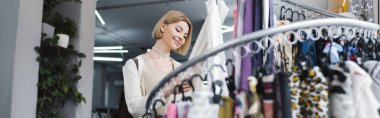 Positive woman touching scarf near rack in second hand, banner 