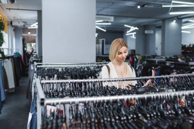 Blonde customer choosing clothes on rack in vintage store 