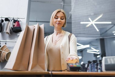 Blonde woman paying with blurred credit card near shopping bags in second hand 