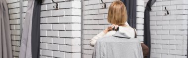 Back view of blonde woman holding hangers near dressing room in second hand, banner 