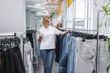 Saleswoman holding digital tablet near jeans on hangers in vintage shop 