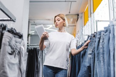 Low angle view of saleswoman looking at digital tablet near jeans in second hand 