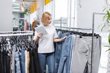 Retailer with digital tablet looking at jeans on rack in second hand  clipart