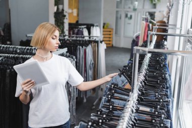 Young retailer holding digital tablet near jeans in vintage store 