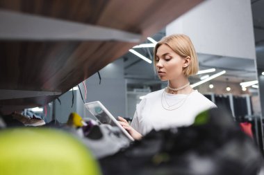 Blonde saleswoman using digital tablet near blurred shelf in second hand  clipart