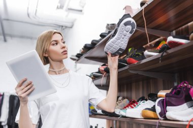 Blonde retailer holding digital tablet and shoe near shelves in second hand 