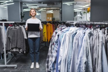 Smiling saleswoman holding empty board near clothes in second hand 