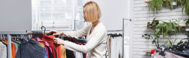 Side view of young customer choosing clothes near plants in second hand, banner 