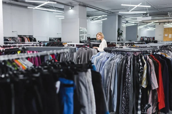 Side view of woman choosing clothes near hangers in vintage shop 