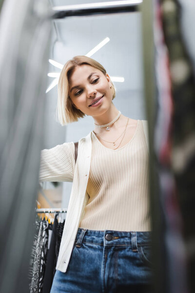 Low angle view of positive woman looking at blurred clothes in second hand 