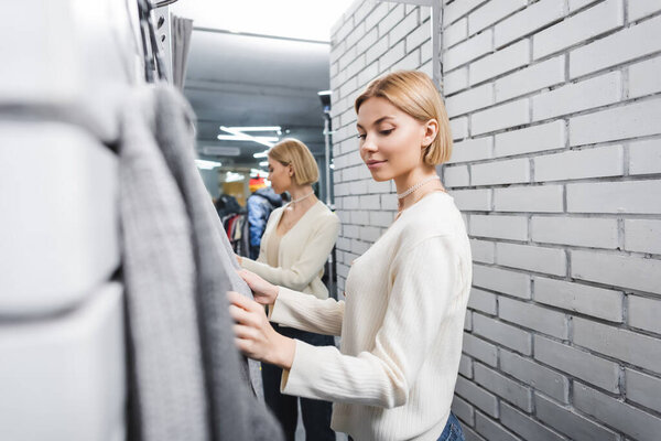 Young customer looking at blurred clothes in dressing room in second hand 