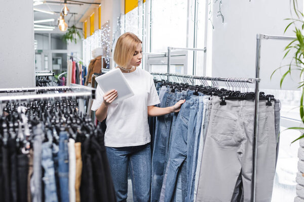 Retailer with digital tablet looking at jeans on rack in second hand 