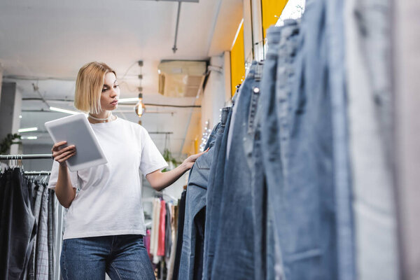 Low angle view of pretty saleswoman looking at jeans and holding digital tablet in second hand 