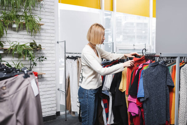 Side view of blonde customer choosing clothes in second hand 
