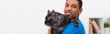 Cheerful african american vet doctor holding maine coon and looking at camera in clinic, banner  clipart