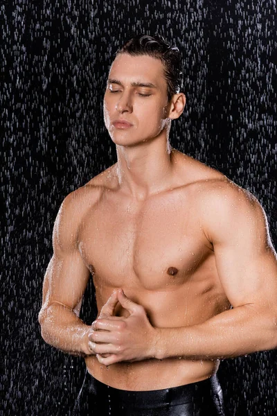 güçlü bir adam kapalı gözlerle poz veriyor ve elleri sımsıkı kapalı siyah arka planda düşen suyun altında.