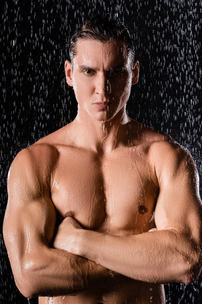 strong man with crossed arms looking at camera under rain shower on black background