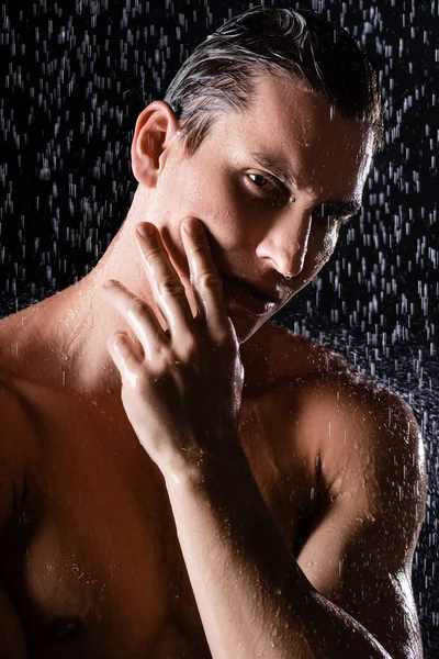 wet man touching face and looking at camera while showering on black background