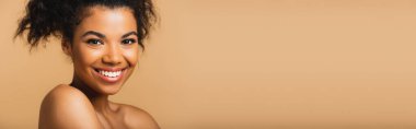 Çıplak omuzları ve bej rengi pankartta izole edilmiş mükemmel bir teni olan neşeli Afro-Amerikan kadın portresi