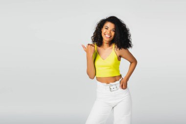 Sarı bluzlu mutlu Afro-Amerikalı kadın. El ele poz veriyor ve baş parmağıyla gri renkte izole edilmiş. 
