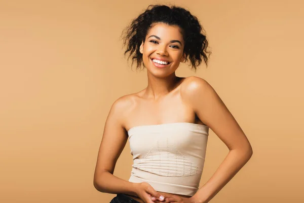 happy african american woman with bare shoulders and natural makeup isolated on beige