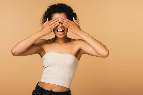 возбужденная африканская американка с голыми плечами, закрывающими глаза руками, изолированными на бежевом 