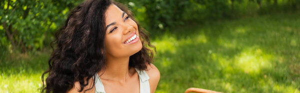 мечтательная африканская американка улыбается и смотрит вдаль в зеленый парк, баннер