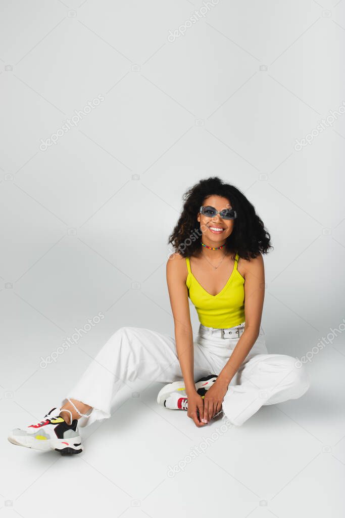joyful african american woman in blue sunglasses and stylish sneakers sitting on grey