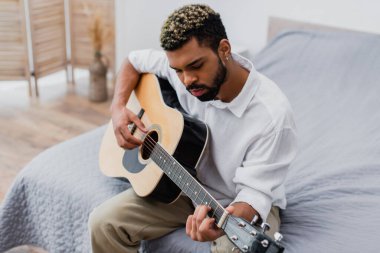 Siyah saçlı ve sakallı genç Afro-Amerikan adam yatak odasında akustik gitar çalıyor.