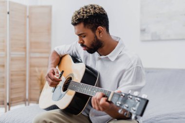 Odaklanmış, saçları boyalı, yatak odasında akustik gitar çalan Afro-Amerikan bir adam.