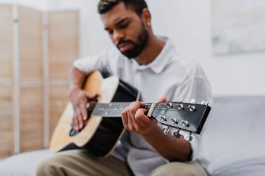 Bulanık, sakallı, evde akustik gitar çalan Afro-Amerikan bir adam.