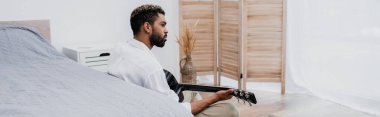 Sakallı ve Afro-Amerikalı genç adam yatağın yanında akustik gitar çalıyor.