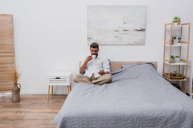 Genç Afro-Amerikalı, boyalı saçlı, elinde kahve fincanı olan ve yatakta dinlenirken akıllı telefon kullanan adam.