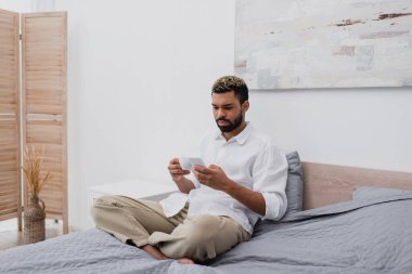 Sakallı Afrikalı Amerikalı bir adam elinde bir fincan kahve tutuyor ve yatakta dinlenirken akıllı telefon kullanıyor.