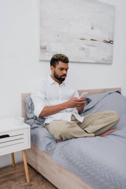 Genç Afro-Amerikalı boyalı saçlı adam yatakta dinlenirken akıllı telefon kullanıyor.