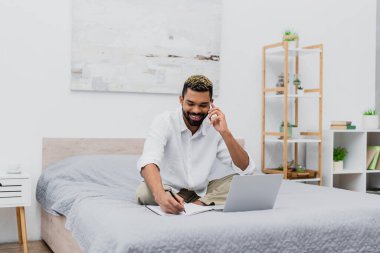 Akıllı telefonla konuşurken gülümseyen ve dizüstü bilgisayarın yanında yazan mutlu Afro-Amerikan adam.