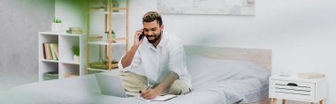 Ön planda bulanık bitki ve pankartla evde çalışırken akıllı telefondan konuşan mutlu Afro-Amerikan adam.